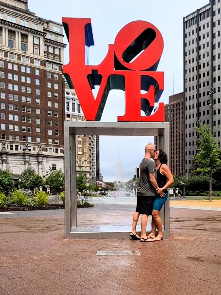James and Kelly pose under the iconic red LOVE sculpture in Philadelphia’s LOVE Park, with city buildings and fountains in the background on a cloudy summer day.
