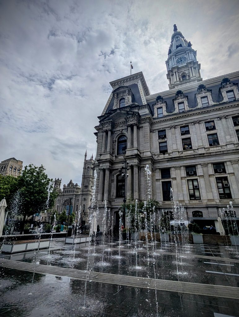 View of Philadelphia City Hall with dramatic clouds and street-level fountains spraying water in the foreground, showcasing the building’s ornate architecture.