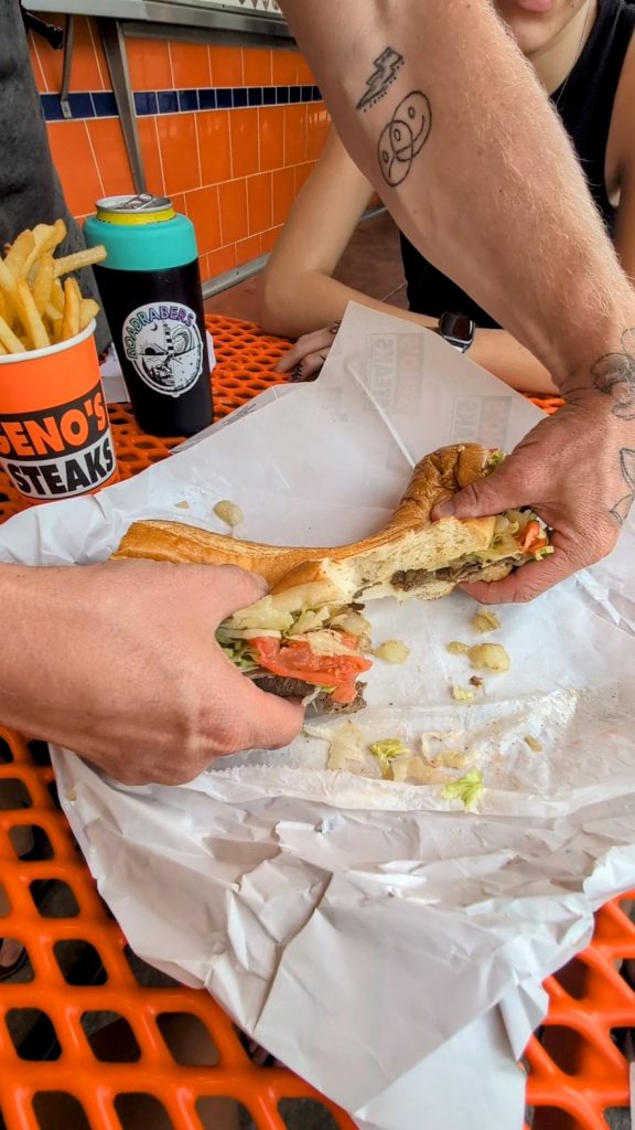 Close-up of a Geno’s Philly cheesesteak being held and eaten at an outdoor table with fries and Geno’s branding visible in the background