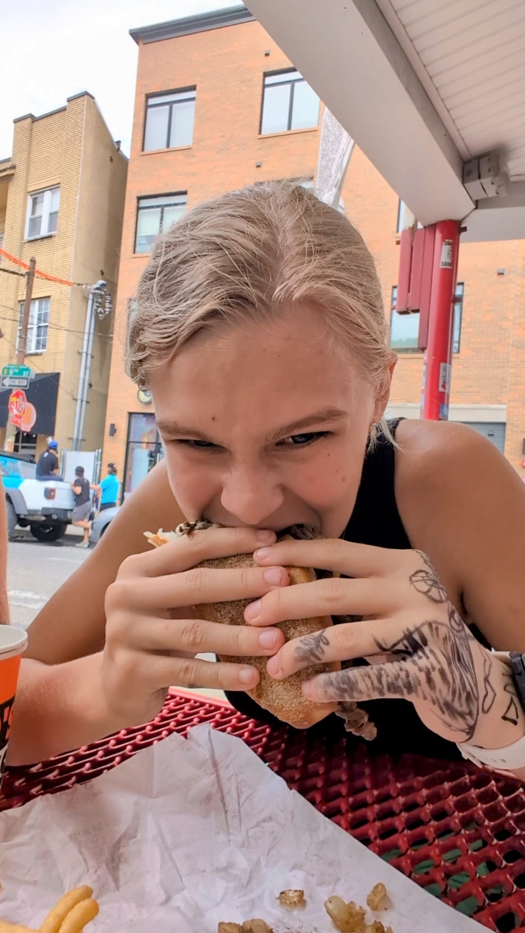 Young girl biting into a Pat’s Philly cheesesteak at an outdoor table with red seating and city buildings in the background.