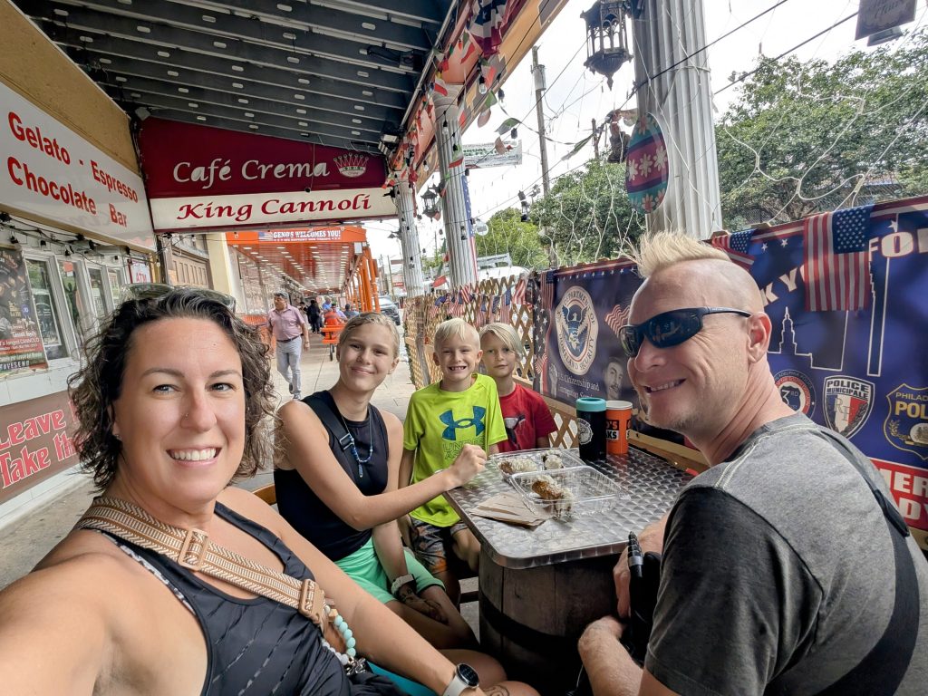 Full-time RV family enjoying pastries together at King Cannoli café in Philadelphia’s Italian Market district, smiling around an outdoor table