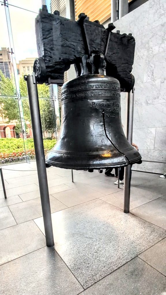 The Liberty Bell on display inside its glass pavilion in Philadelphia, showcasing the iconic crack and historic inscription with natural light coming through the windows.