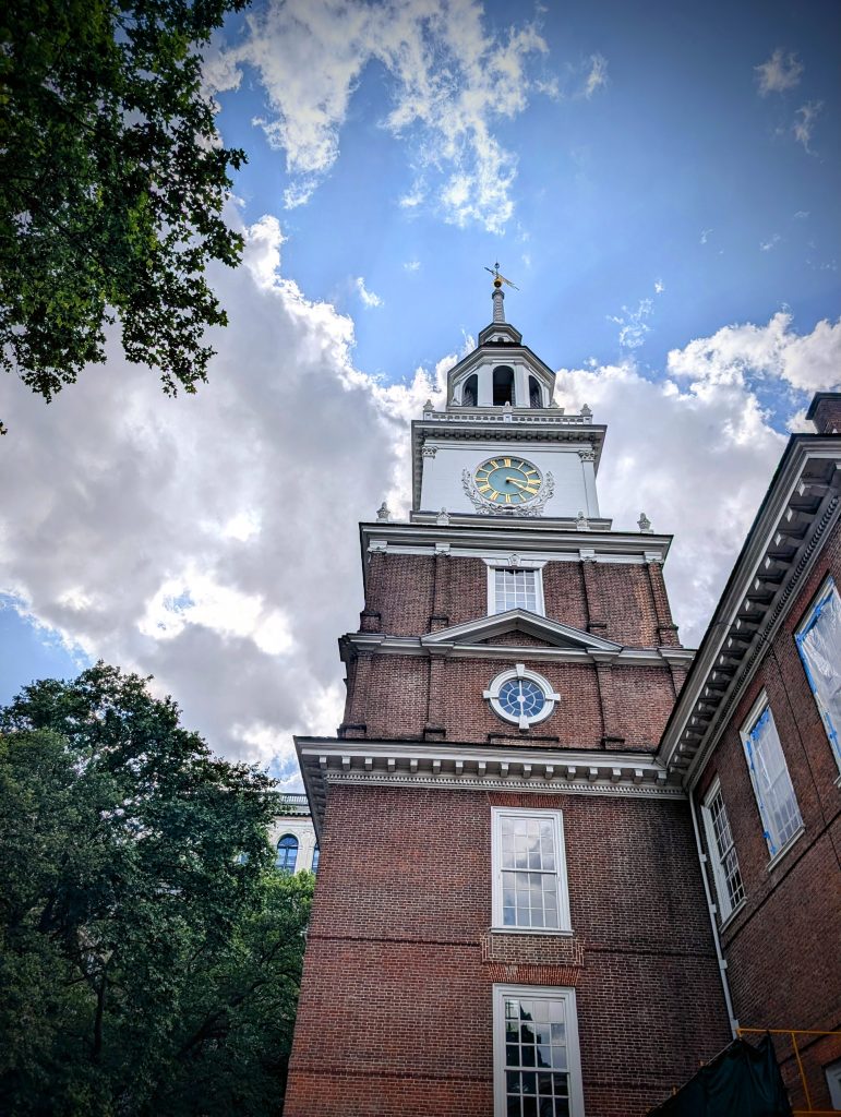 Independence Hall in Philadelphia under a dramatic sky, with its iconic clock tower framed by leafy green trees.