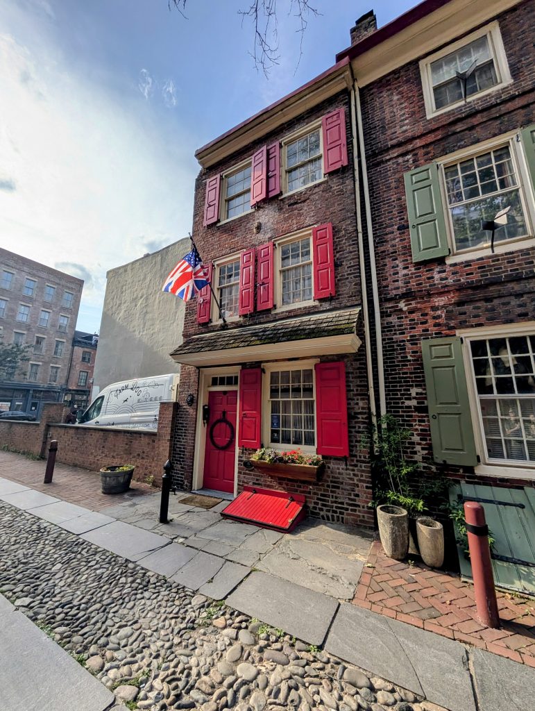 Charming red-shuttered brick home for sale on historic Elfreth’s Alley in Philadelphia, with cobblestone street and American and British flags displayed.