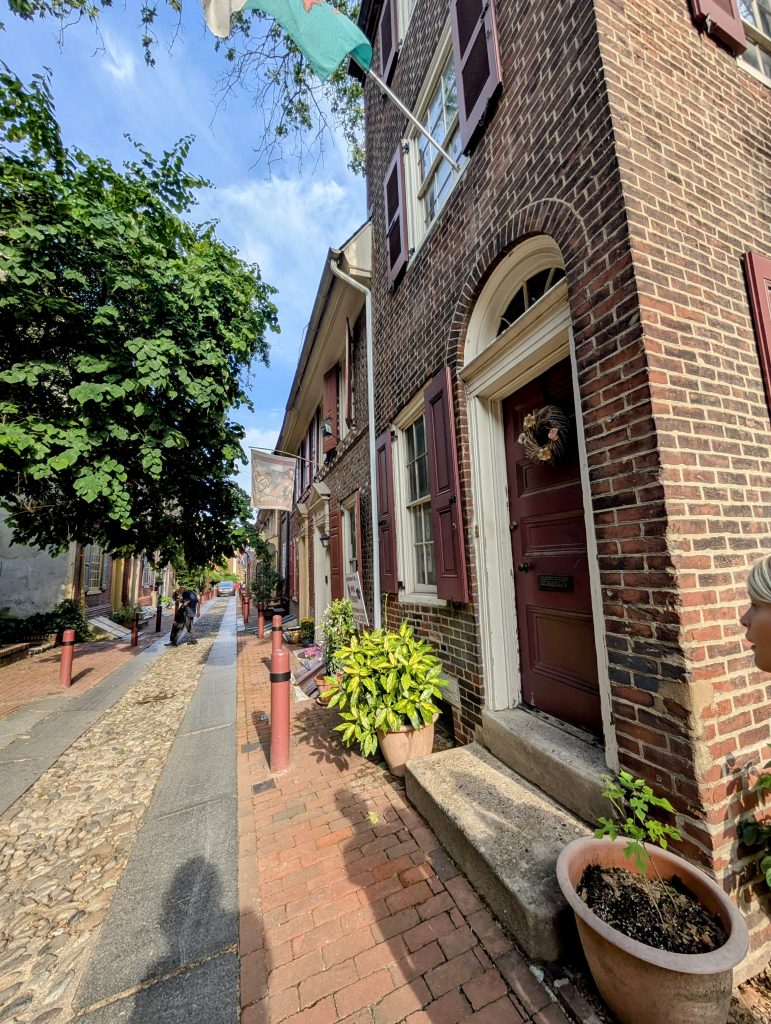 Brick homes lining the cobblestone path of Elfreth’s Alley in Philadelphia, with flower pots and colonial-style doorways on a sunny day.